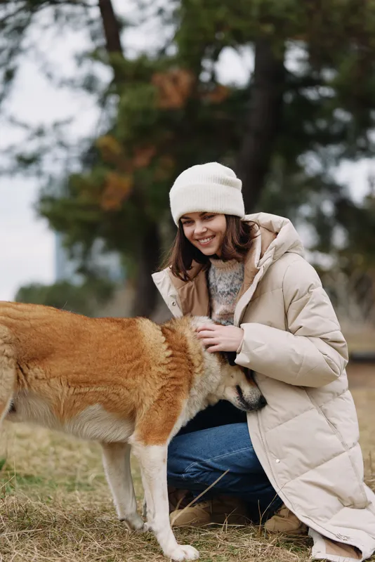 A woman in a warm jacket and beanie kneels in a park outdoors and gently pets a dog, capturing a candid winter moment with natural light, cozy attire, and a calm affectionate companionship in nature
