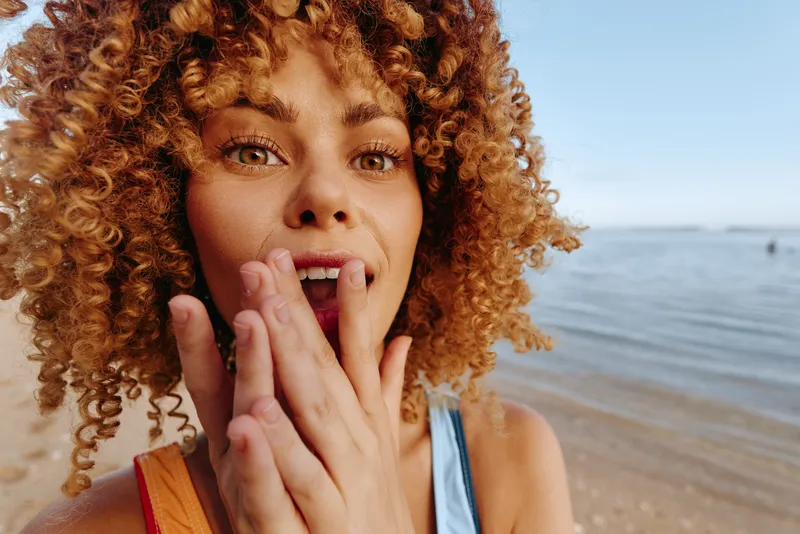 A young woman with curly hair expresses surprise and joy while standing on a sandy beach near calm water under a clear sky during daytime.