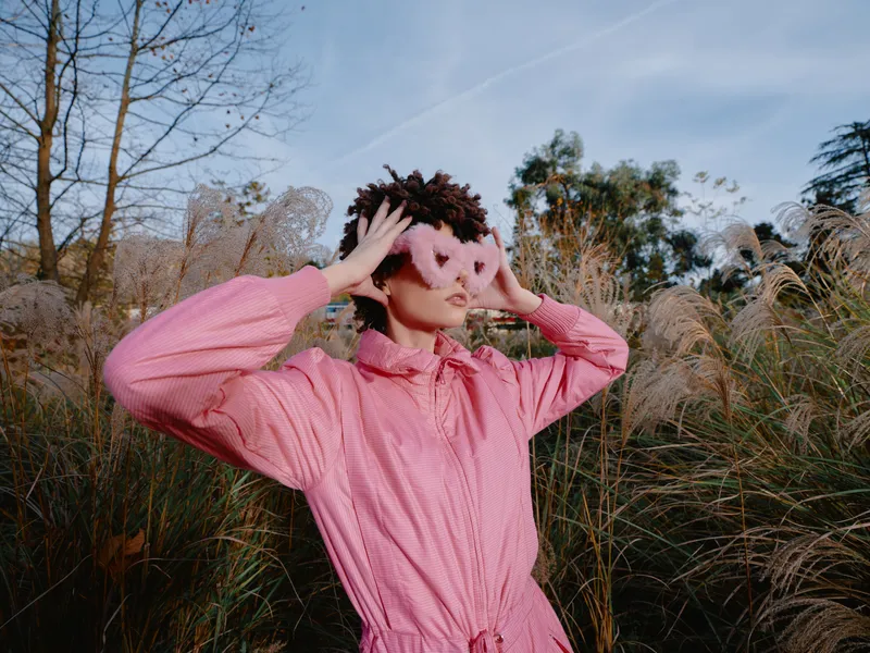 Portrait of a person in a pink outfit adjusting a playful mask in tall grass outdoors under a clear sky, showcasing creative fashion and bold nature photography with a carefree stance against a