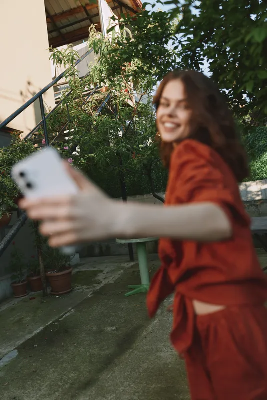 Young woman in a red dress takes a candid selfie outdoors, smiling at her phone with lush greenery and a sunlit courtyard backdrop, a relaxed lifestyle moment