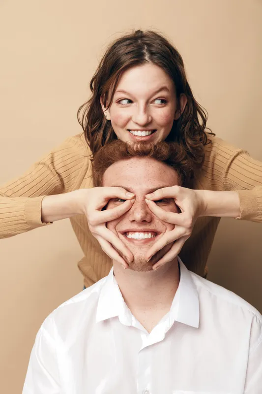 Happy young woman playfully covers eyes of smiling man with her hands forming glasses shape. Casual clothing and beige background create warm, fun, and intimate couple moment concept.