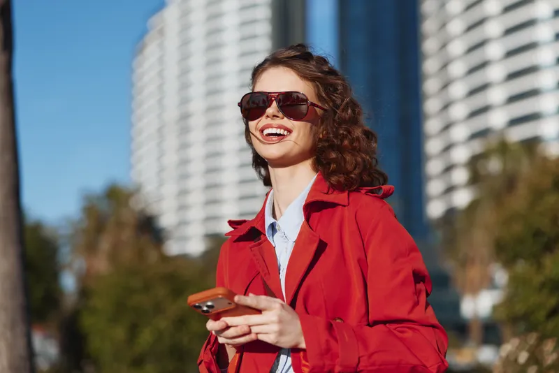 Cheerful woman outdoors, smiling with sunglasses on, red coat fluttering, urban backdrop with skyscrapers, bright daylight, carefree pose, candid fashion moment