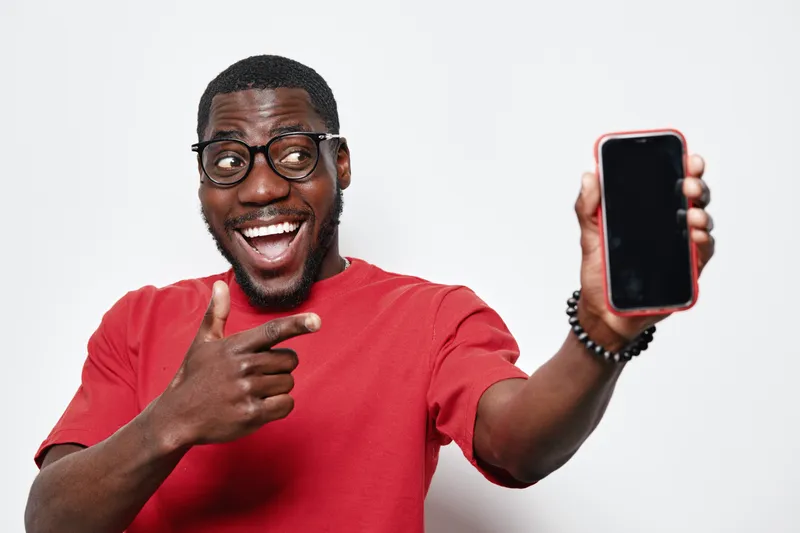 African American man with dark skin and glasses smiling and pointing at smartphone in red shirt on white background. Happy young male showing mobile phone screen.