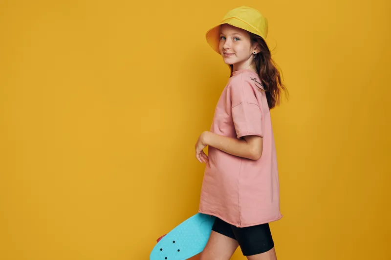Smiling girl in yellow hat holding blue skateboard on yellow background. Side view portrait in casual summer outfit.
