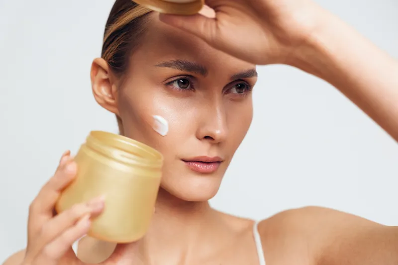 Young woman applies cream from a jar on her cheek, concentrating as a simple skincare routine unfolds in a bright studio, conveying calm self care and focus.