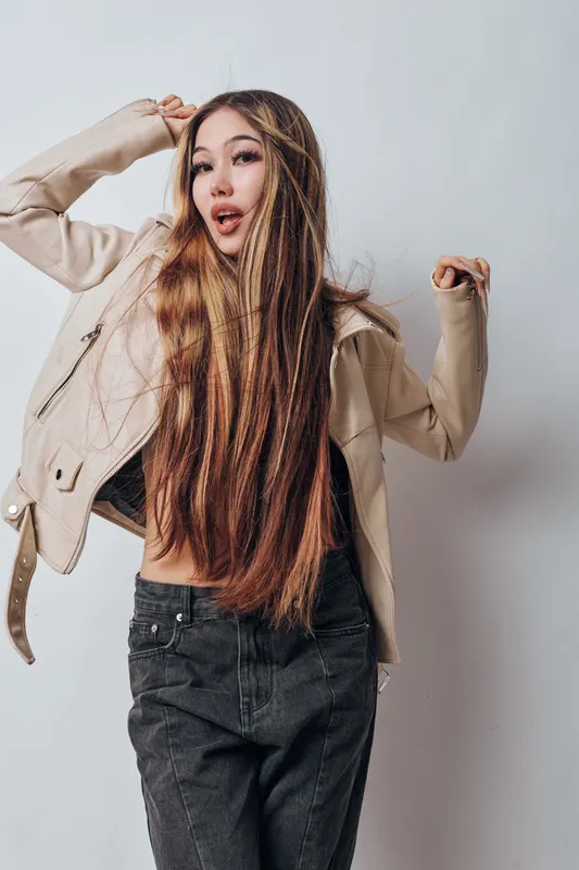 Young woman with long hair wearing beige jacket and black jeans posing against white background with confident expression and casual style in studio photography session.