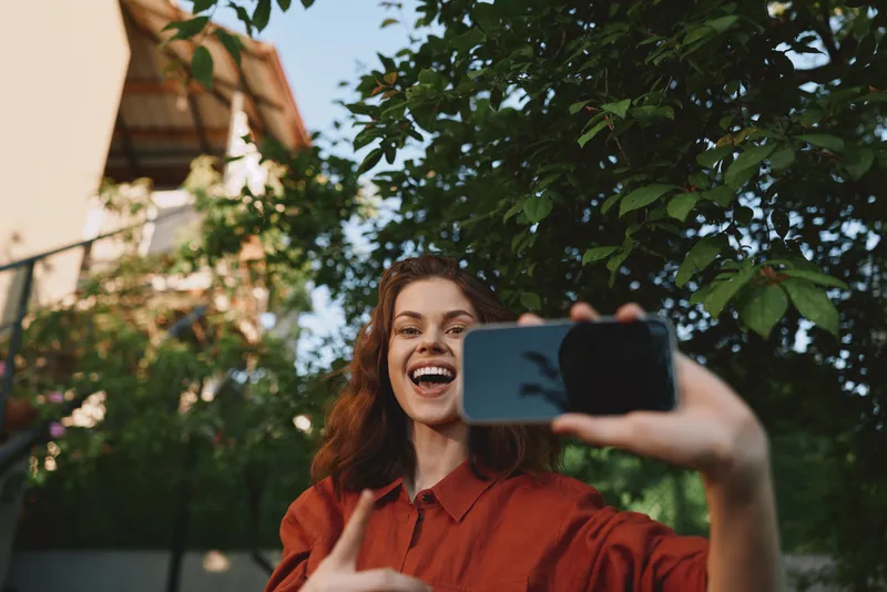 Selfie taking woman in a red shirt smiles in an outdoor garden, capturing a candid lifestyle moment