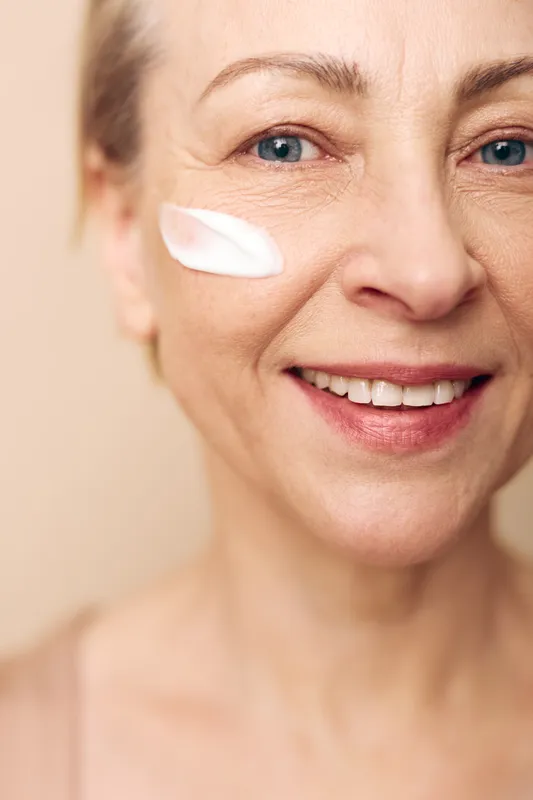 Close-up of smiling mature woman with blue eyes applying white face cream on cheek. Skincare concept for aging skin, beauty routine, hydration, and natural healthy glow.
