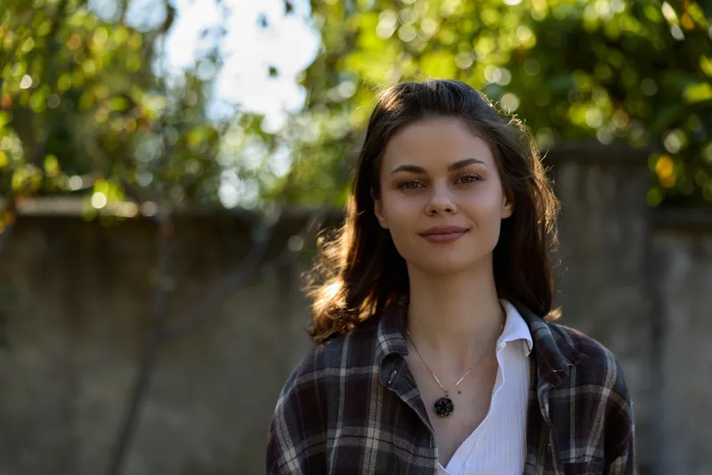 Young woman with shoulder length hair stands outdoors in a plaid shirt relaxed and confident as soft sunlight filters through leafy trees