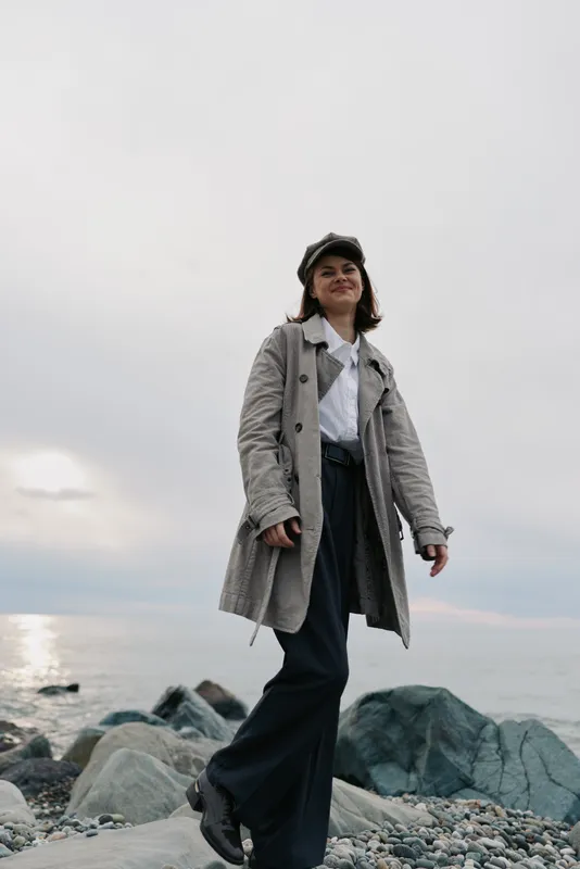 Young woman in a long coat and hat walks confidently on rocky beach near calm sea under cloudy sky. Casual fashion and outdoor lifestyle concept with natural scenery and relaxed mood.