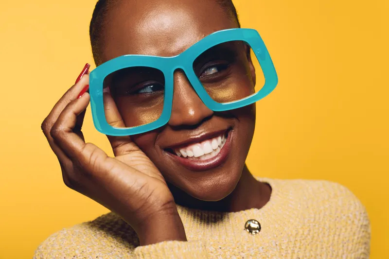 Close-up of a smiling black woman with dark skin wearing large blue glasses and a beige sweater. Bright yellow background highlights her joyful expression and stylish accessories.