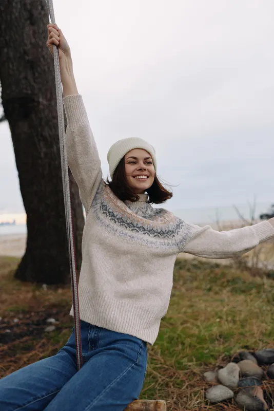 Woman on a swingset outdoors, smiling and relaxed, wearing a cozy sweater and beanie, enjoying fresh air and nature on a breezy day by the park