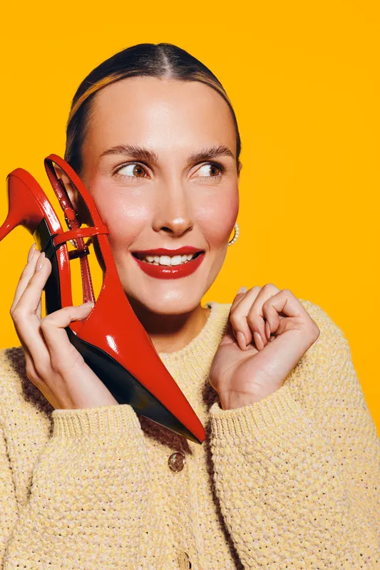 Young woman with red high heel shoes holds them near her face smiling. Bright yellow background and cozy beige sweater create cheerful and stylish fashion concept with vibrant colors.