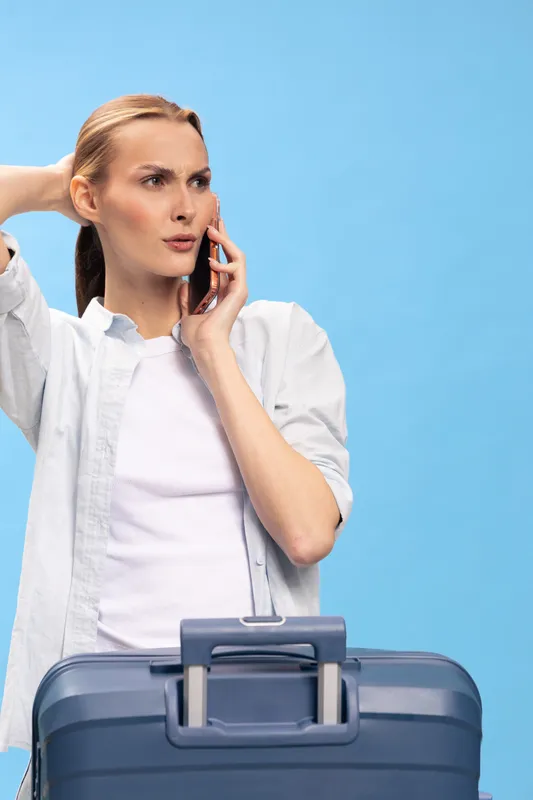 Young woman with suitcase talks on phone, looking concerned. She wears casual white shirt and stands against blue background. Travel, communication, and planning concept.