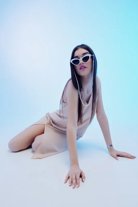 Young woman beauty fashion model poses on the floor wearing stylish sunglasses and a beige dress. Studio shot with soft blue lighting. Concept for elegance and modern style.