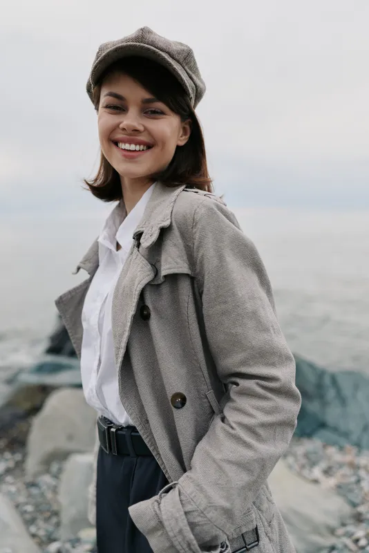 Smiling young woman in casual beige coat and cap stands near rocky shore. She wears white shirt and black trousers, enjoying outdoor fresh air with a joyful, confident expression.