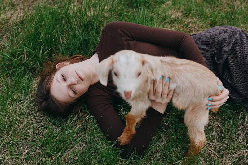A girl lies on grass, holding a goat kid in a gentle embrace in bright natural light. A calm outdoor moment shows affection, connection, and care in a simple lifestyle scene.