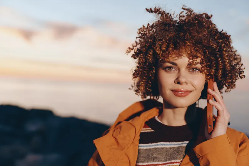 Woman with curly hair outdoors, smiling gently and posing in an autumn landscape. She adjusts her hair near her cheek, wearing a knitted sweater. Warm sunset light over mountains.