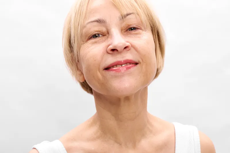 Elderly woman portrait, smiling with gentle expression, blonde short hair, studio closeup against clean white background, soft even light, calm mood, healthy skin