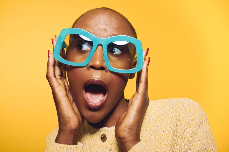 Surprised black woman with dark skin wearing large blue glasses and a cream sweater on yellow background. Concept of fashion, expression, and colorful style with female model.