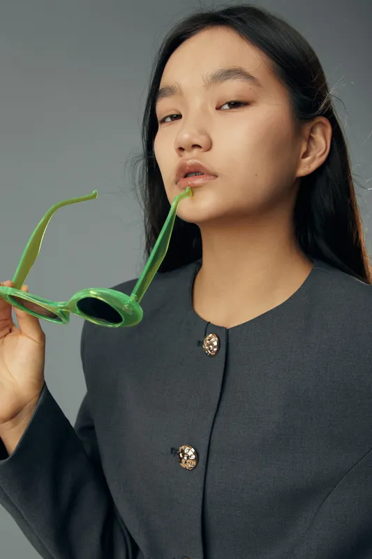 Portrait of confident young woman holding green sunglasses near lips wearing dark gray jacket with gold buttons, posing in studio with neutral background, stylish and modern fashion look.
