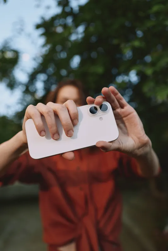 Smiling woman outdoors holds a smartphone for a selfie, blurred green background, casual lifestyle moment, candid photography, natural light, carefree mood and joyful connection with nature
