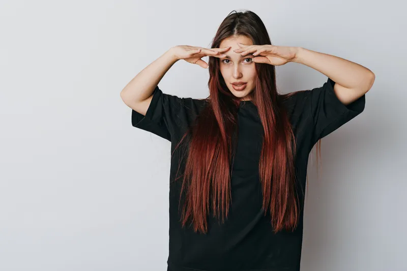 Young woman with long red hair and black t shirt looking forward with hands above eyes on white background for vision, focus and searching concept in studio portrait.