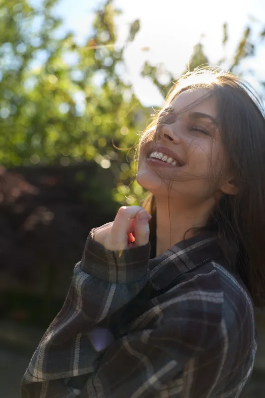 A relaxed woman in a plaid shirt enjoys sunny outdoor warmth, smiling with eyes closed. A candid lifestyle moment capturing happiness, confidence, and simple daily joy under bright light today