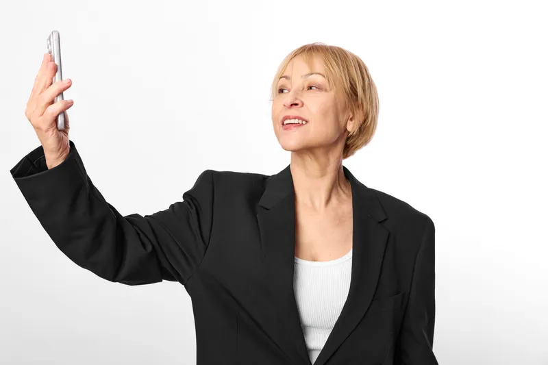Woman in business suit takes selfie with smartphone, phone in hand, studio portrait, smiling, white background, confident pose, clean copy space