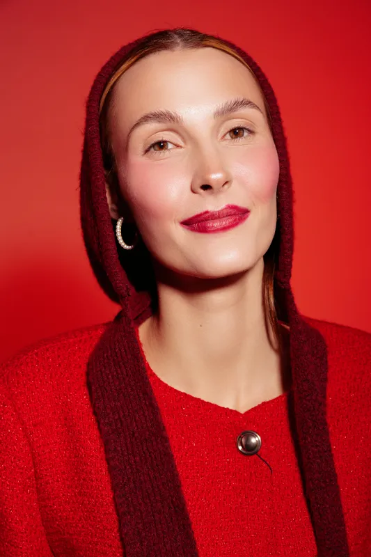 Portrait of a confident woman with natural makeup wearing a red sweater and hood against a red background. Concept of fashion, beauty, and minimalistic style with warm tones.