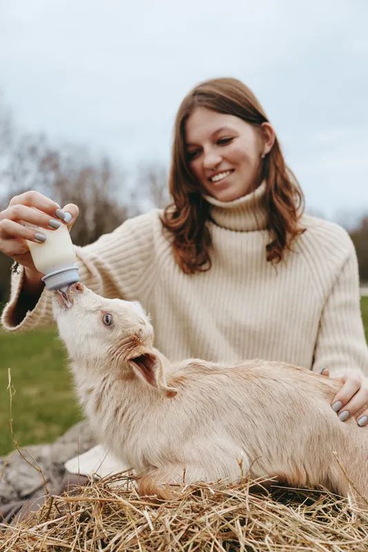 Young woman in beige sweater feeds a baby goat with a bottle outdoors on a cloudy day, showing a gentle connection and caring moment in a natural lifestyle setting.