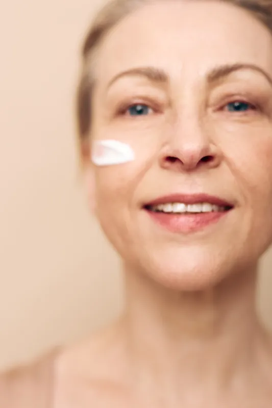Mature woman with short hair applies white face cream on cheek, smiling gently on beige background. Concept of skincare, aging care, beauty routine, and self-care.