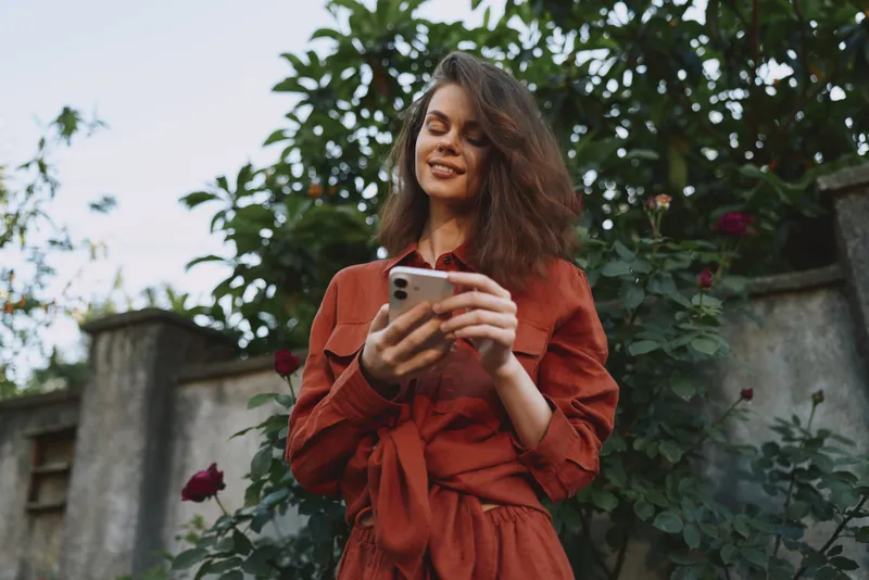 Cheerful woman outdoors in a red dress, smiling at her phone, sunlit garden moment, relaxed pose, soft breeze, vibrant colors, casual lifestyle scene