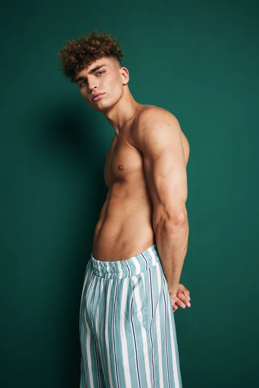 Young man with curly hair posing shirtless in striped pants against green background, showing muscular build and confident expression in studio portrait.