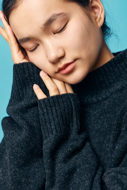 Young woman with closed eyes wearing black knitted sweater resting her face on palm against blue background. Relaxed person with folded hands showing calm expression and minimal makeup.