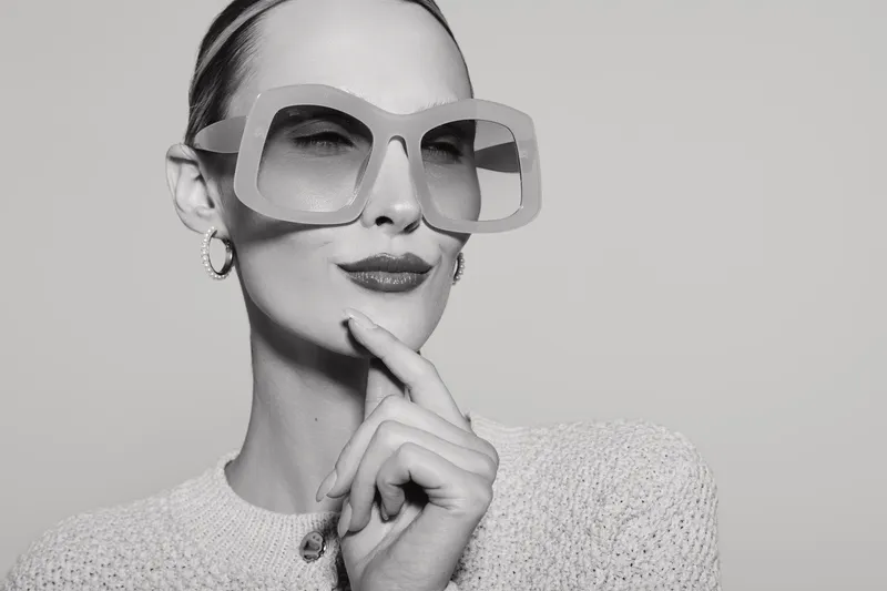 Elegant white woman with pale skin and slicked back hair wears oversized glasses and textured sweater while posing thoughtfully against plain background. Concept of style and fashion.