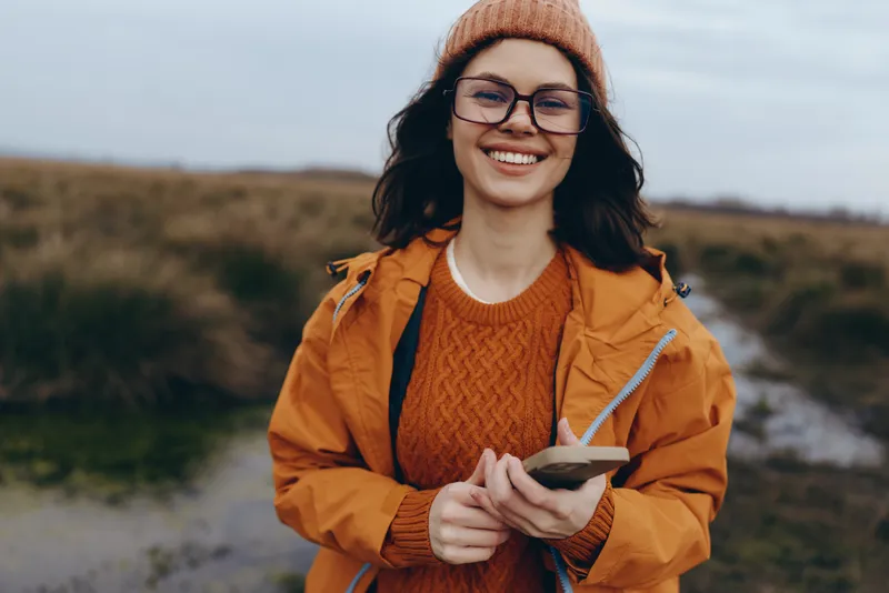 Woman lifestyle portrait outdoors, smiling through glasses, in orange jacket and beanie, holding a smartphone. Autumn countryside hills blur behind her, modern communication happy mood.