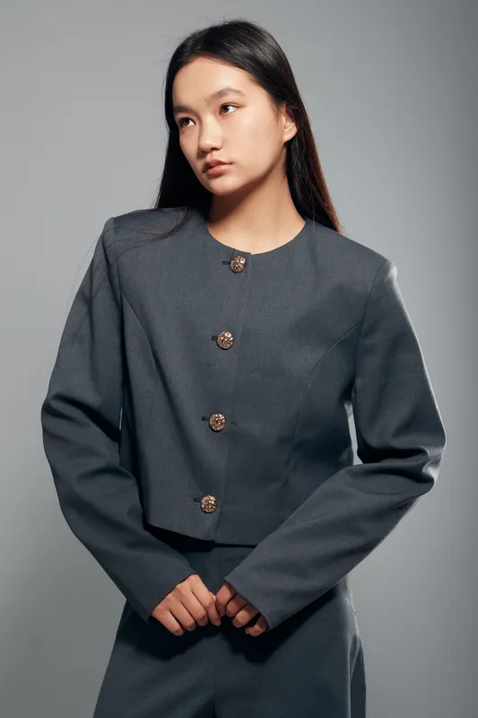 Young woman wearing gray formal suit with decorative buttons posing isolated on gray background, looking aside with thoughtful expression and hands clasped in front.