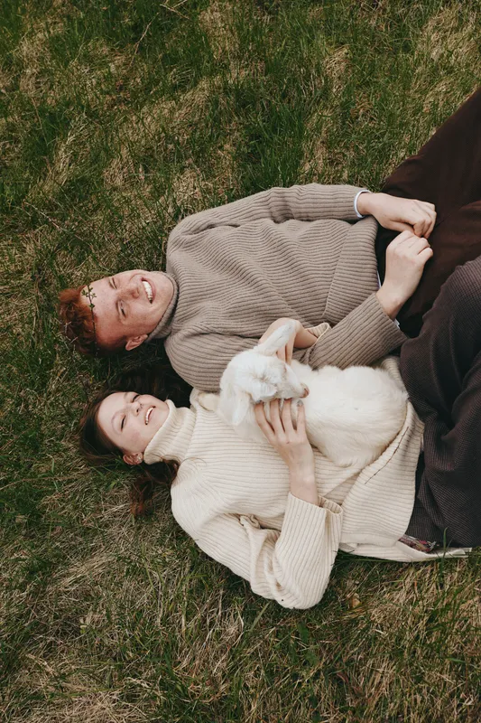 Couple lying on grass in cozy sweaters, playful goat kid between them, sunlit outdoor moment, relaxed embrace, natural colors, rural lifestyle portrait. Soft evening light, gentle breeze.
