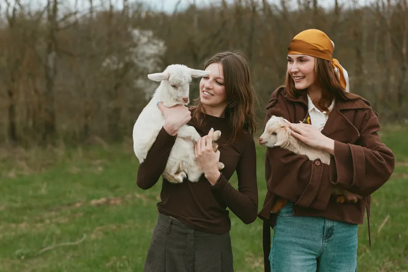 Two smiling women in casual clothing hold fluffy goat kids in a grassy field, enjoying a rustic outdoor moment, friendly interaction, candid lifestyle scene with playful animals in daylight.