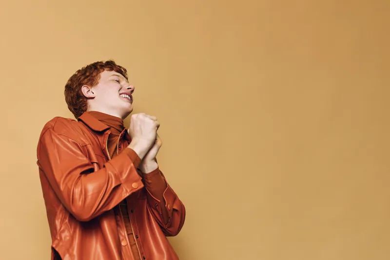 Young man wearing brown leather jacket with closed eyes and clasped hands showing excitement or hope on beige background, casual style portrait indoors.