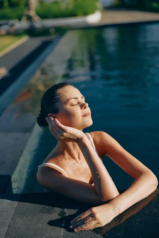 Relaxed woman by a sunlit pool, eyes closed, gentle breeze, soft reflections on water, carefree posture, natural colors, outdoor lifestyle moment, capturing a tranquil, sunlit scene of casual comfort