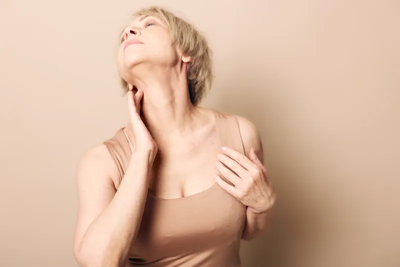 Mature woman with short blonde hair touches her neck and chest wearing beige tank top. Soft lighting highlights skin texture and natural aging. Concept of self-care and aging gracefully.