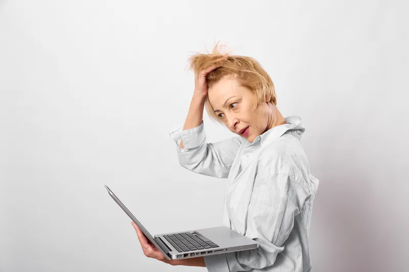Elderly woman with laptop showing fatigue, holding head with headache, tech stress mood, studio portrait on white background, indoor home office desk concept
