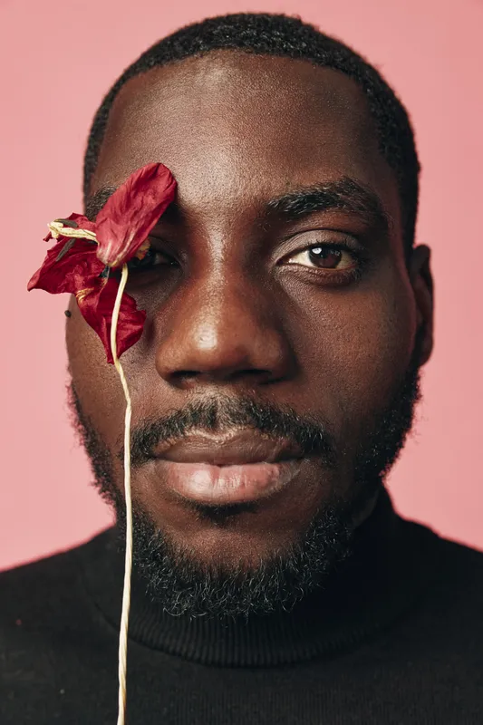 African man with dark skin and beard holding dried red flower near eye in creative conceptual visual style on pink background. Portrait with artistic expression and fashion mood.