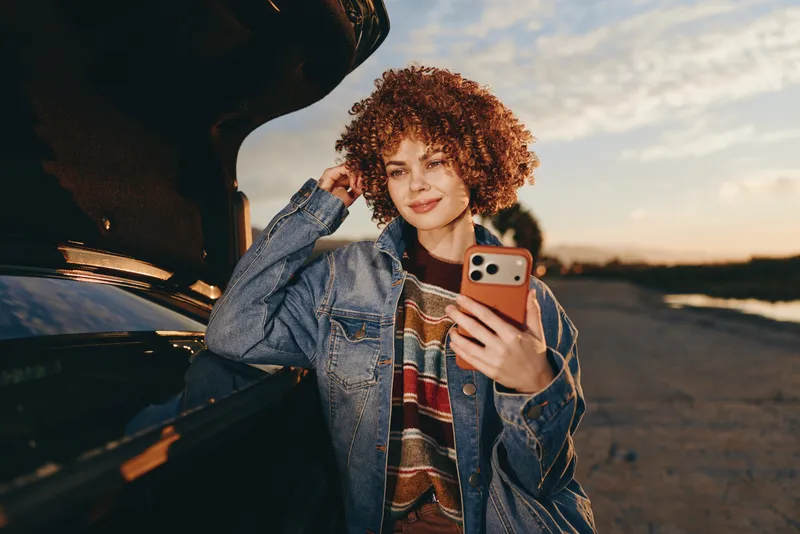 Woman selfie outdoors by a car at sunset, curly hair and denim jacket, holding smartphone near open door, smiling, warm golden light, travel lifestyle