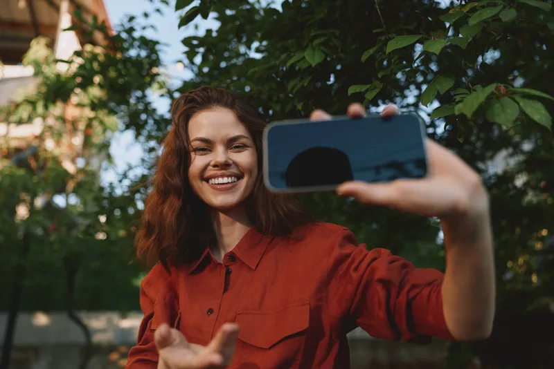 Cheerful woman outdoors, smiling while taking a selfie with a smartphone, red shirt, lush greenery background, natural light, carefree lifestyle moment