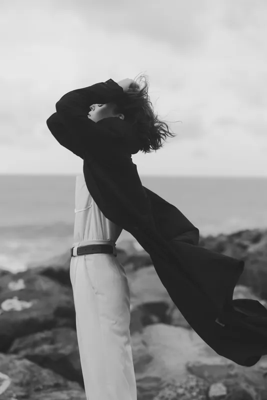 Windy coastal frame with a figure lifting a scarf toward the horizon, black and white mood emphasizing motion, freedom and stark contrast.