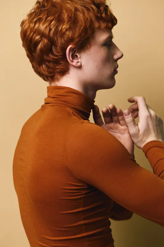 Profile of young man with red curly hair wearing brown turtleneck adjusting sleeve in studio with plain beige background. Closeup of side face and hand movements for clothing.
