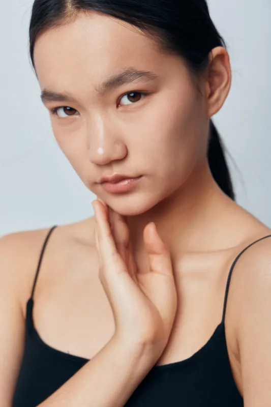 Portrait of young woman with natural skin and soft expression touching her neck indoors. Beauty closeup with minimal makeup, healthy face, and casual black top on light background.